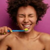 Everyday life, morning routine and teeth cleaning concept. Horizontal shot of shirtless dark skinned woman brushes teeth with tooth brush, stands naked against purple wall, being in good mood