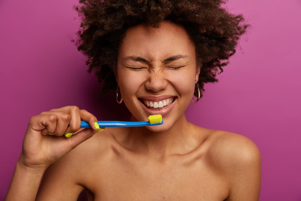 Everyday life, morning routine and teeth cleaning concept. Horizontal shot of shirtless dark skinned woman brushes teeth with tooth brush, stands naked against purple wall, being in good mood