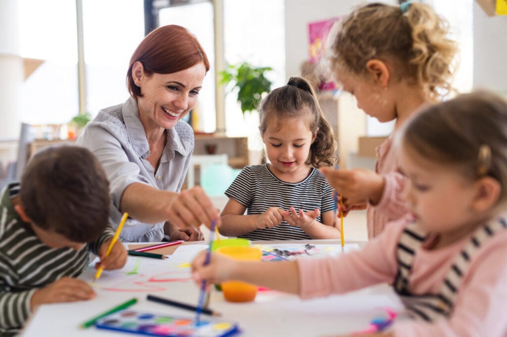 Group of small nursery school children with teacher indoors in classroom, painting.