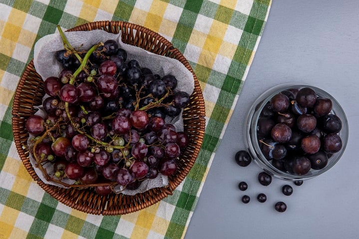 top view of black grape in basket on plaid cloth and grape berries in bowl on gray background